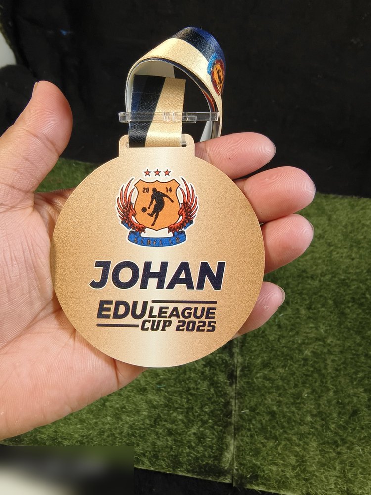 Bahagian BELAKANG medal Edu League Cup 2025 — permukaan emas penuh dengan logo pasukan, teks besar 'JOHAN' dan 'EDU LEAGUE CUP 2025'. Contoh medal 2 sided untuk kedudukan pemenang.