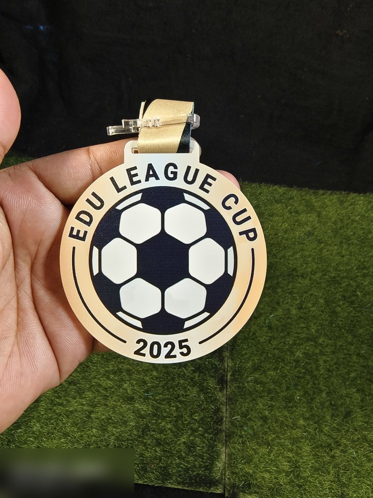 Medal Edu League Cup 2025 — liga bola sepak. Bentuk bulat dengan bola sepak besar di tengah (putih dan biru navy) dan bingkai emas. Teks 'EDU LEAGUE CUP 2025' melengkung. Reben emas. Design ringkas da