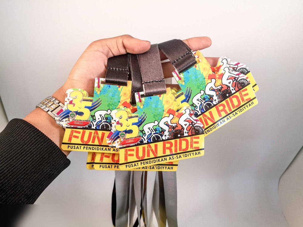 Medal Fun Ride Pusat Pendidikan As-Sa'idiyyah. Bentuk custom dgn sepanduk KUNING besar bertulis 'FUN RIDE', silhouette pelumba basikal warna-warni dan latar hijau-biru. Tab kulit coklat. Gambar studio
