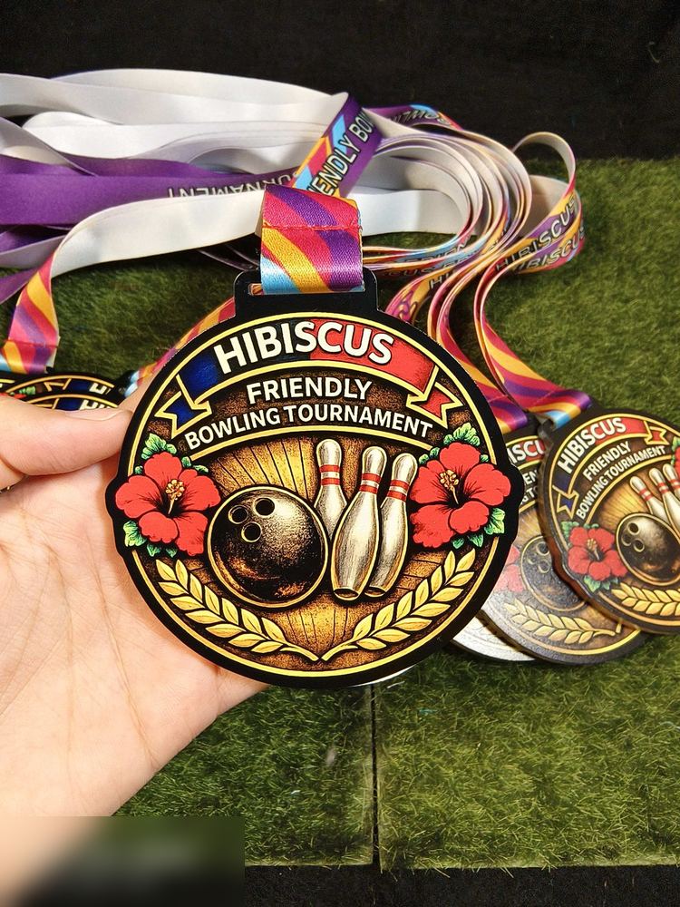 Medal Hibiscus Friendly Bowling Tournament 2026. Bentuk bulat dengan bingkai emas berkilat, latar hitam, bola dan pin bowling di tengah, bunga raya merah di kiri kanan dan kalungan padi emas. Nampak s