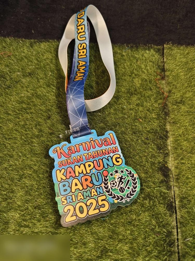 Medal Karnival Sukan Tahunan Kampung Baru Sri Aman 2025. Bentuk custom mengikut susunan teks berwarna-warni, dengan kalungan daun dan ikon sukan (bola sepak, pelari, badminton) di sebelah kanan. Warna