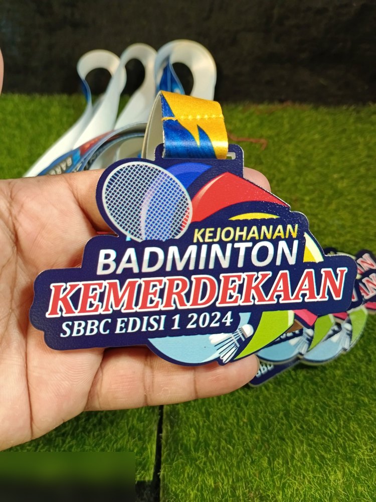 Medal Kejohanan Badminton Kemerdekaan SBBC Edisi 1 2024. Bentuk custom, latar biru navy dgn raket besar dan bentuk geometri warna-warni (merah, kuning, hijau, biru). Teks 'KEMERDEKAAN' merah menonjol.