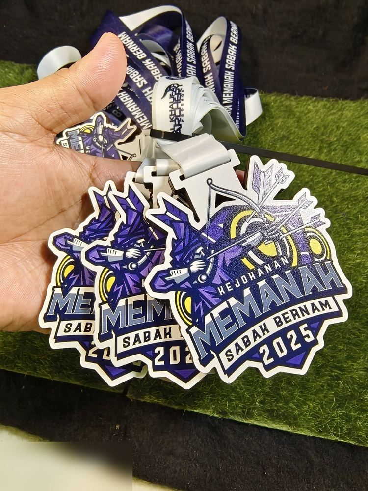 Medal Kejohanan Memanah Sabak Bernam 2025. Bentuk custom dgn pemanah gaya kristal/low-poly warna UNGU dan biru, bulatan kuning di belakang. Lanyard ungu-putih bercetak. Versi 2025 (bandingkan dgn vers