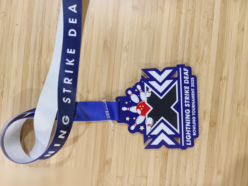 Medal Lightning Strike Deaf Bowling Tournament 2025 — kejohanan komuniti PEKAK/OKU. Bentuk custom seperti anak panah chevron bertindih dgn 'X' besar di tengah, warna biru royal dan putih, pin bowling