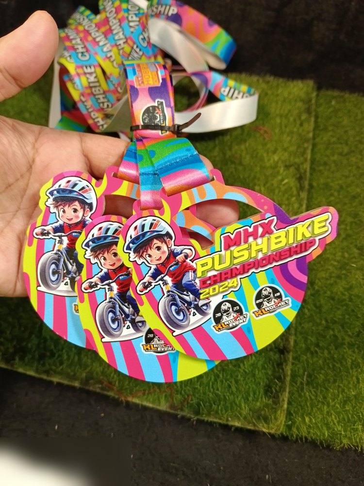Medal MHX Pushbike Championship 2024. Bentuk custom bulat dgn watak KARTUN budak menunggang pushbike. Warna paling terang dlm koleksi — merah jambu, kuning, biru, hijau berjalur. Lanyard pelangi. Sang