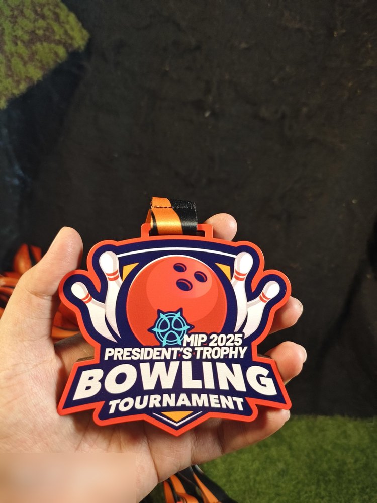 Medal MIP 2025 President's Trophy Bowling Tournament. Bentuk custom dgn bola bowling OREN besar di tengah dan pin putih, latar biru navy dgn bingkai oren. Reben oren-hitam. Versi 2025 (bandingkan dgn