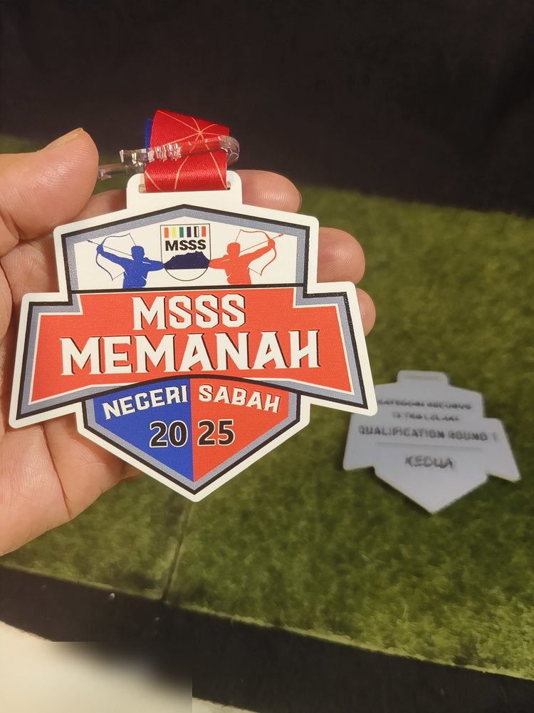 Medal MSSS Memanah Negeri Sabah 2025. Bentuk perisai dgn peta Sabah di tengah, warna merah, biru dan putih, dua pemanah di atas. Gambar tunjuk SEKALI bahagian BELAKANG (putih berfros) yang bertulis ka
