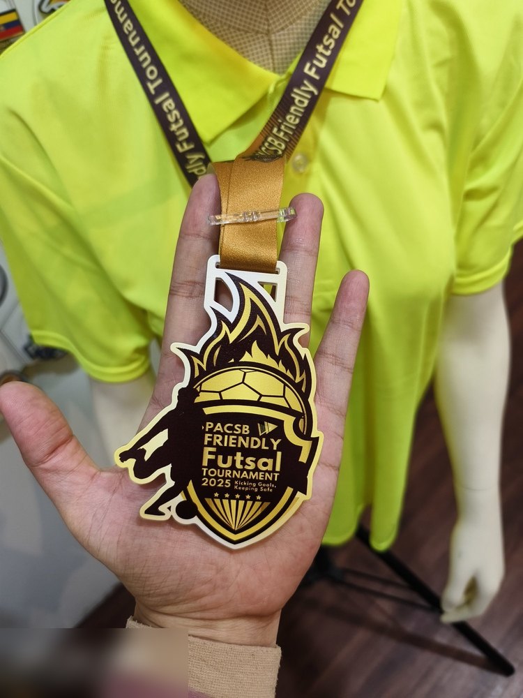 Medal PACSB Friendly Futsal Tournament 2025. Bentuk custom perisai dengan bola sepak berapi dan silhouette serigala/harimau. Warna coklat marun dan emas — nampak premium. Moto 'Kicking Goals, Keeping