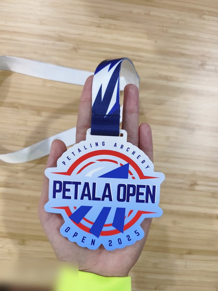 Medal Petala Open 2025 Petaling Archery dipegang di tangan atas latar meja kayu — tunjuk saiz sebenar berbanding tapak tangan. Warna putih, biru royal dan merah. Lanyard biru-putih corak kilat.