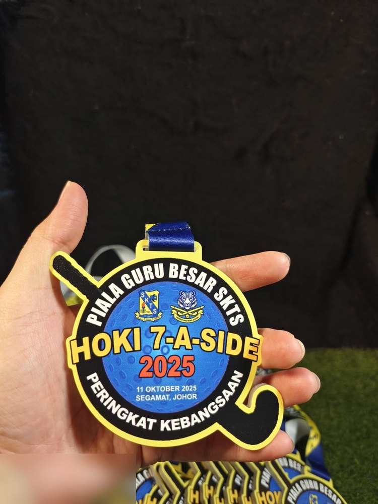 Medal Hoki 7-A-Side Piala Guru Besar SKTS 2025, Peringkat Kebangsaan, Segamat Johor. Bentuk custom mengikut kayu hoki melengkung. Warna hitam, kuning dan biru dengan tekstur bola hoki. Reben biru gela