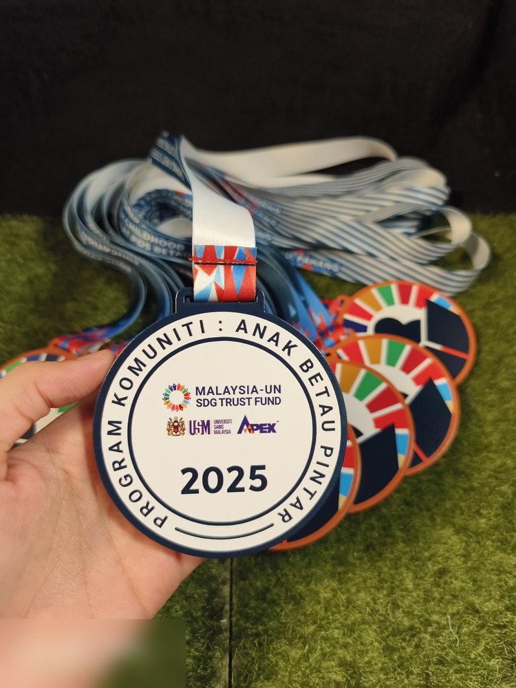Medal Program Komuniti: Anak Betau Pintar 2025, Malaysia-UN SDG Trust Fund bersama USM dan APEX. Bentuk bulat, muka depan putih dengan bingkai biru navy dan logo penaja. Muka belakang tunjuk roda warn