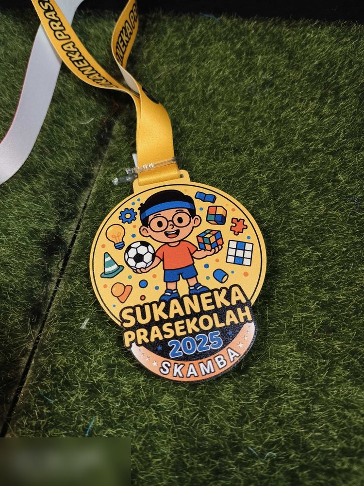 Medal Sukaneka Prasekolah SKAMBA 2025. Bentuk bulat warna kuning dengan ilustrasi kartun budak berkaca mata memegang bola sepak dan rubik, dikelilingi ikon mainan. Teks 'SUKANEKA PRASEKOLAH 2025 SKAMB