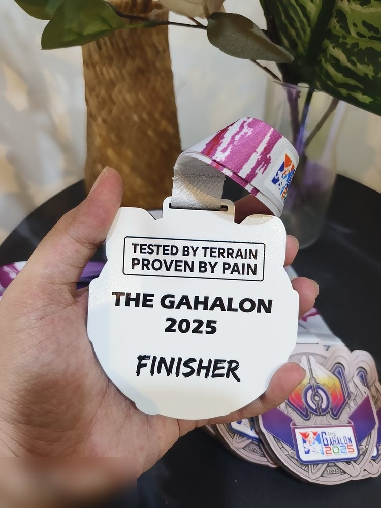 Bahagian BELAKANG medal The Gahalon 2025 — putih bersih dengan teks 'TESTED BY TERRAIN, PROVEN BY PAIN', 'THE GAHALON 2025' dan 'FINISHER'. Lanyard corak merah jambu. Contoh medal 2 sided di mana bela