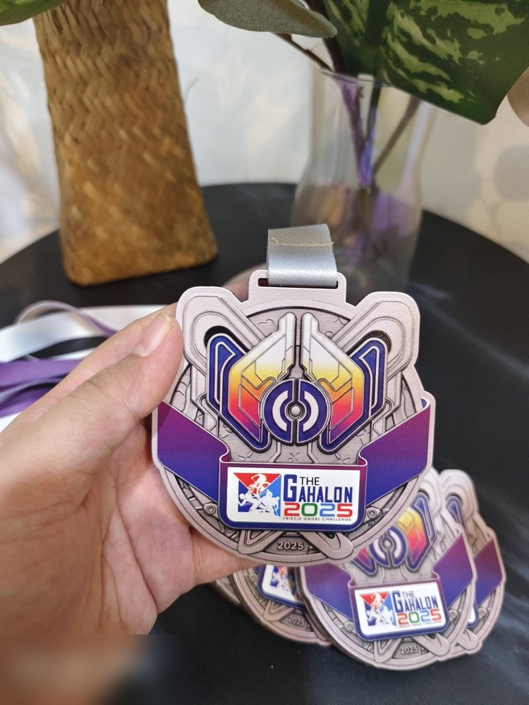 Medal The Gahalon 2025 — Mieco Ganai Challenge. Bentuk custom besar bergaya futuristik/mekanikal dengan bingkai perak dan pita ungu-oren gradasi. Logo berwarna di tengah. Reben perak. Design paling ko
